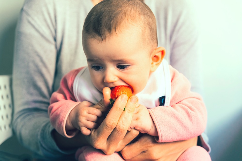 Bebé muy pequeño chupando una fresa que sostiene su madre, antes de empezar con el BLW